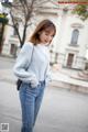 A woman in a gray sweater and jeans is standing on the street.