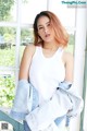 A woman in a white tank top and denim jacket leaning against a window.